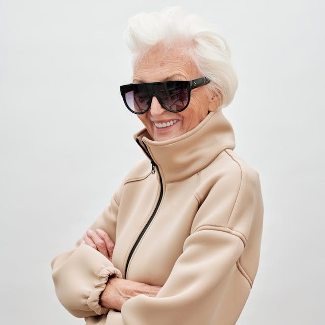 femme souriante très âgée aux cheveux entièrement blancs avec une paire de lunettes noires très moderne et une veste sportswear moderne aussi